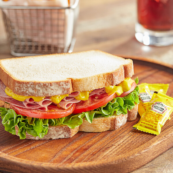 A sandwich with meat and vegetables on a wooden plate with a Heinz Yellow Mustard packet.