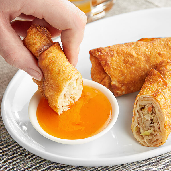 A hand dipping a fried egg roll into a small bowl of orange Kraft Sweet'n Sour Sauce.