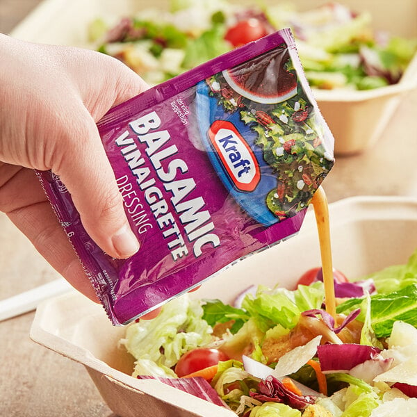 A hand pouring a purple Kraft Balsamic Vinaigrette packet onto a salad.