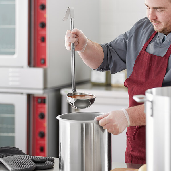 A man in a red apron using a Choice stainless steel ladle to stir a pot.