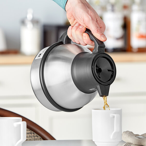 A person pouring coffee from a Bunn stainless steel coffee carafe into a white mug.