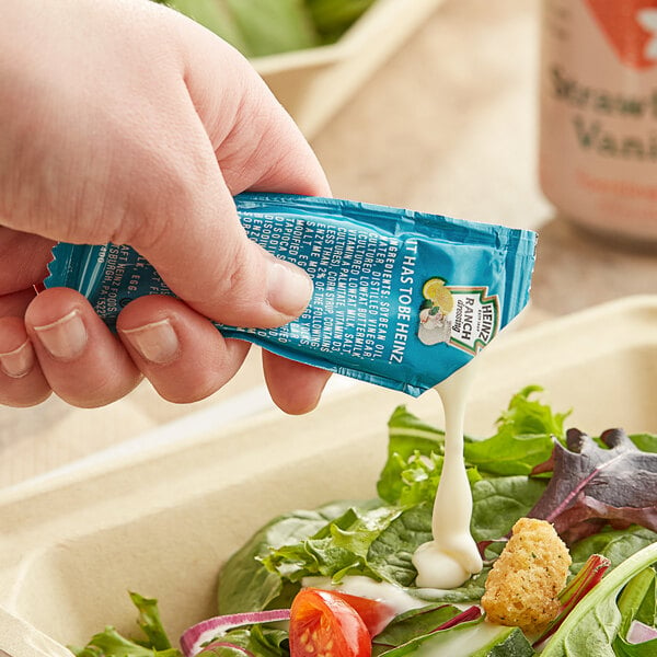 A hand holding a blue Heinz Ranch dressing packet over a salad