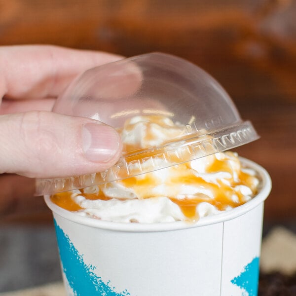 A hand holding a Solo clear plastic lid over a cup of ice cream with whipped cream and caramel sauce.