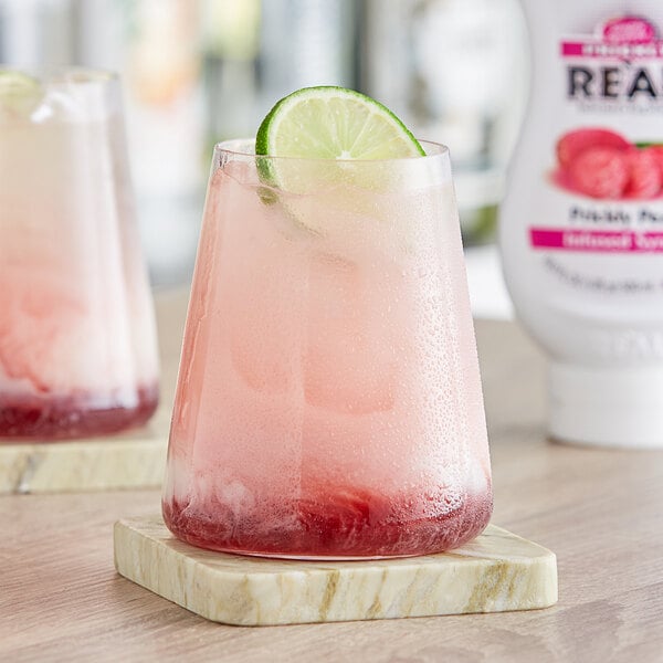 A glass of pink liquid with a lime wedge on top next to a bottle of Real Prickly Pear Puree Infused Syrup.