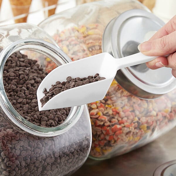 A hand holding a white Choice flat bottom scoop over a jar of chocolate chips.