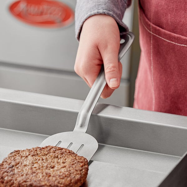 A person using a Choice stainless steel slotted spatula to flip a burger.