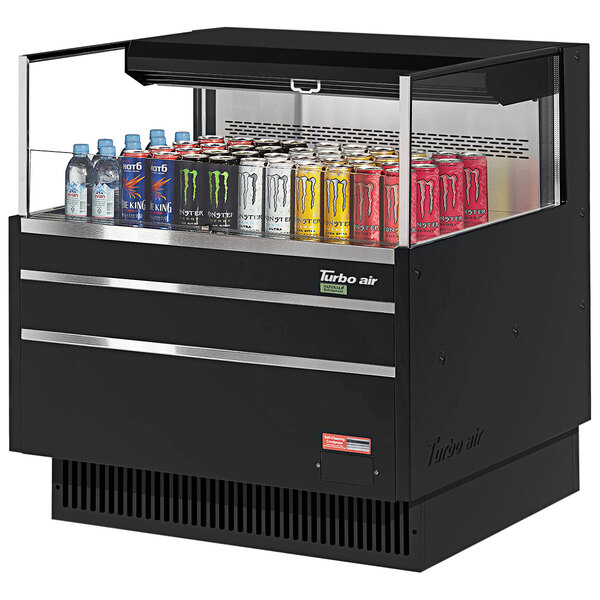 A black Turbo Air horizontal refrigerated open curtain merchandiser on a counter with cans of soda and different colored cans inside.