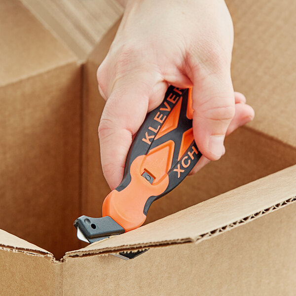 A hand using an orange and black Klever Kutter X-Change to cut a box.