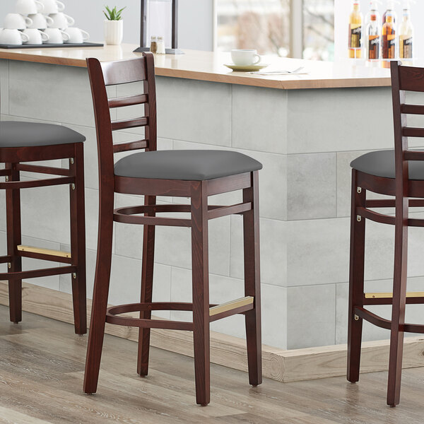 Three Lancaster Table & Seating mahogany wood bar stools with dark gray vinyl seats on a wooden counter.