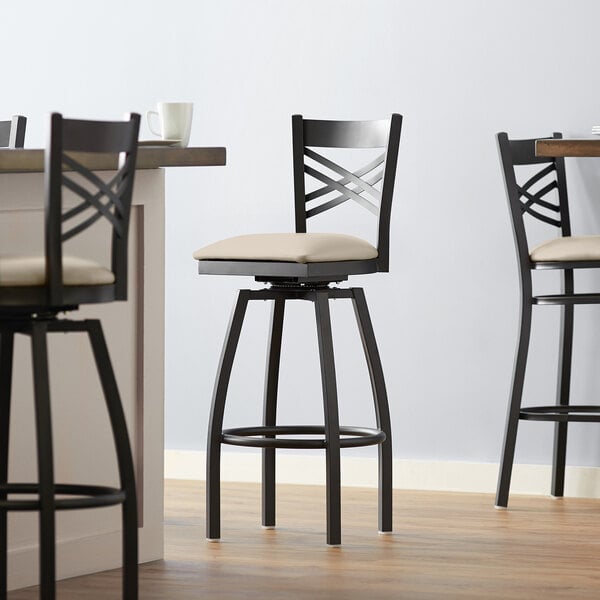 A close up of a Lancaster Table & Seating black finish cross back swivel bar stool with a light gray vinyl padded seat.