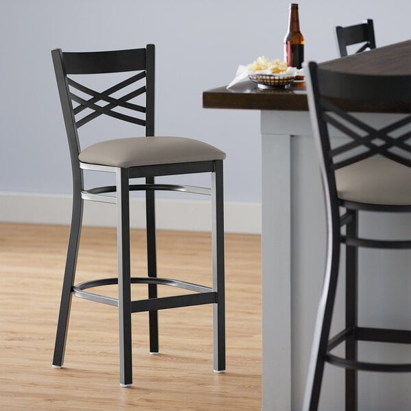 A Lancaster Table & Seating black cross back bar stool with a dark gray cushion on the seat.