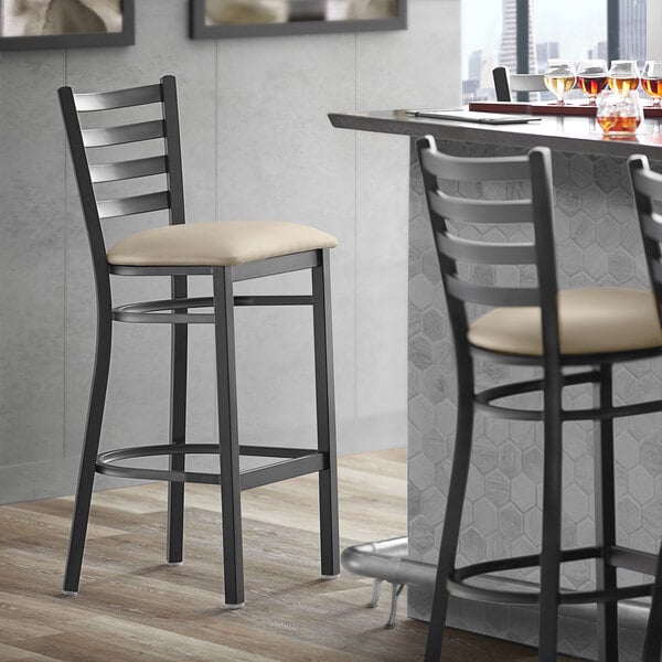 A Lancaster Table & Seating black finish ladder back bar stool with a light gray vinyl padded seat next to a table.