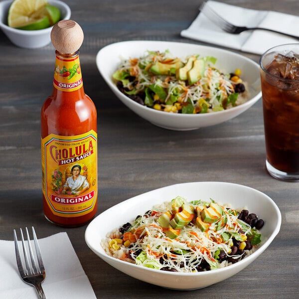 A bowl of salad with avocado and cheese on a table with a bottle of Cholula Original Hot Sauce.