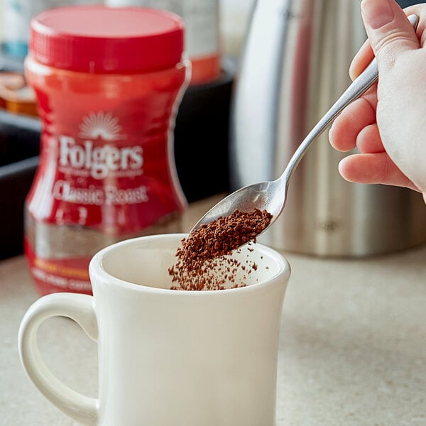 A person pouring Folgers Classic Roast Instant Coffee into a white mug.