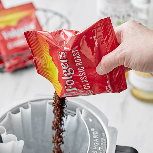 A hand pouring Folgers Classic Roast coffee from a red packet into a coffee machine.