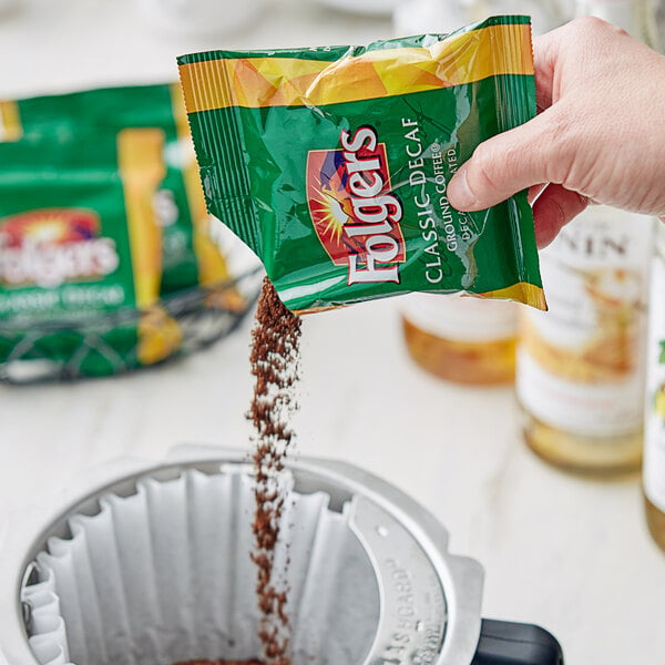 A hand pouring Folgers Classic Decaf coffee from a green and yellow packet.