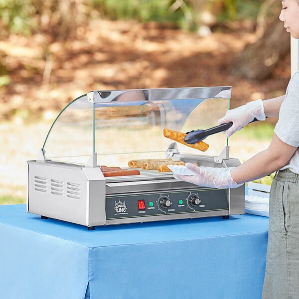 A woman using a Carnival King hot dog roller to prepare hot dogs at an outdoor event.
