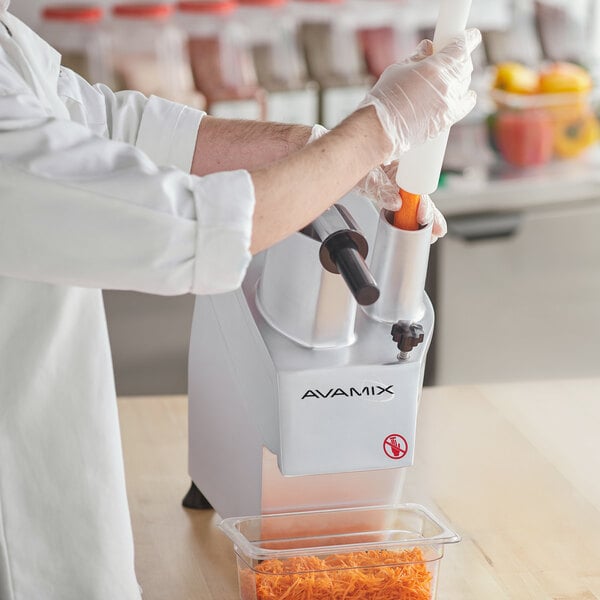 A person in a white coat and gloves uses the AvaMix food processor to shred carrots into a plastic container.