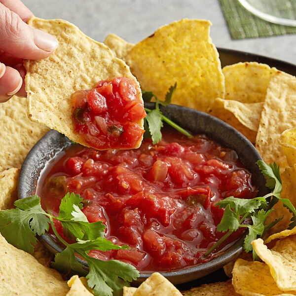 A hand holding a chip with Del Sol Red Mexican Salsa over a bowl of salsa on a table with more chips.