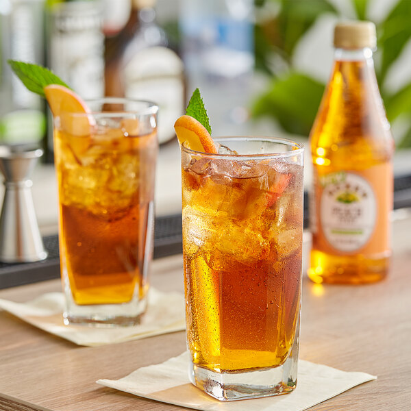 A bottle of Rose's Peach Syrup on a table with a glass of peach iced tea and mint.