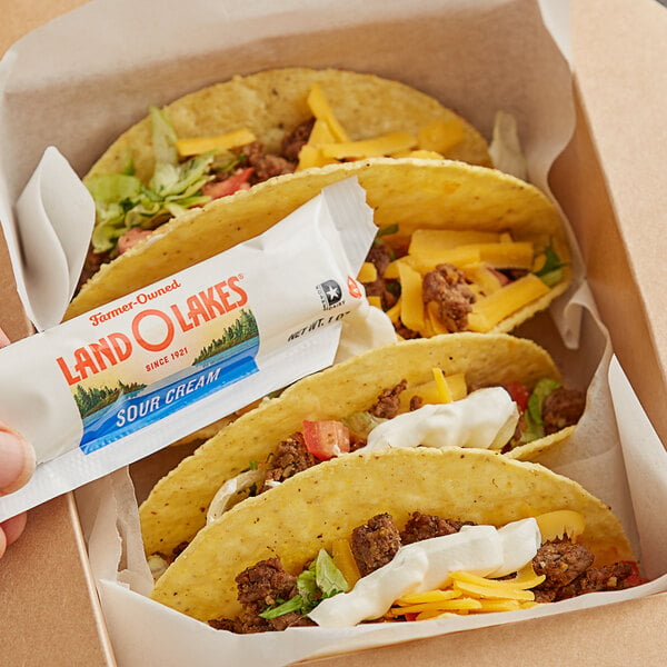A hand holding a Land O Lakes sour cream packet over a box of tacos with cheese and meat.