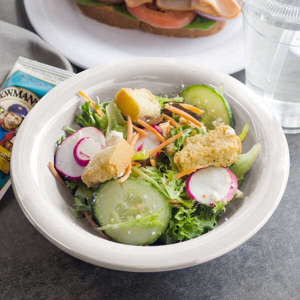 A bowl of salad with a sandwich and a glass of water on a counter with a Carlisle bone rimmed melamine bowl of salad.