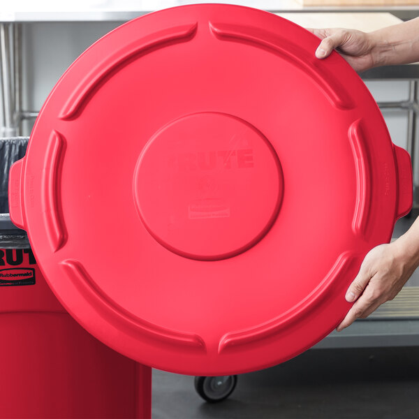 A person holding a red Rubbermaid BRUTE trash can lid.