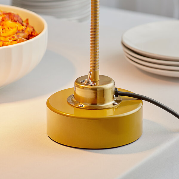 An Avantco gold weighted base for a bulb warmer heat lamp on a table with a bowl of pasta.