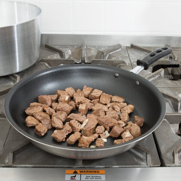 A Vollrath stainless steel frying pan with meat cooking on a stove top.