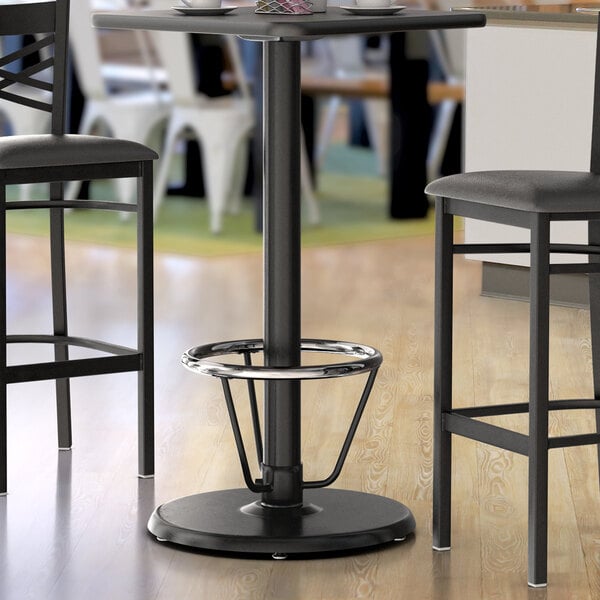 A Lancaster Table & Seating black bar height table base with a foot ring on a table with a cup on top and two chairs.