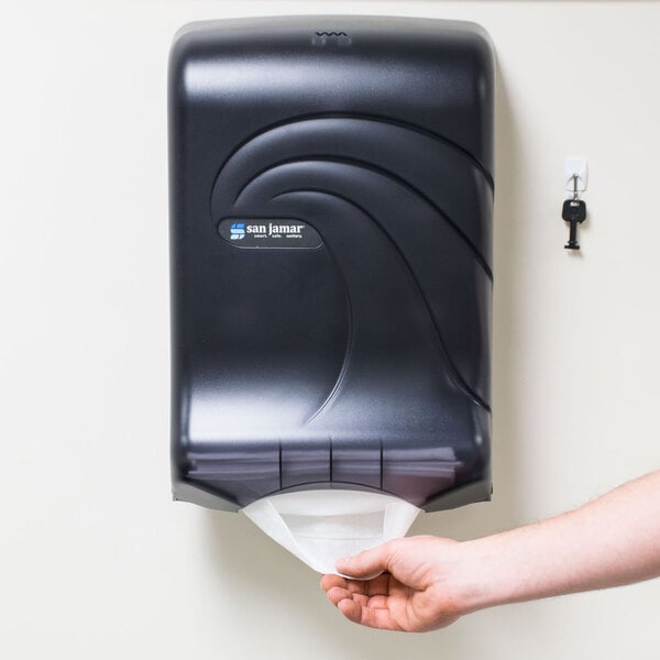 A person's hand using a black San Jamar paper towel dispenser.