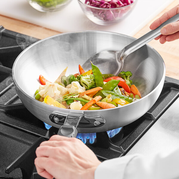 A person cooking vegetables in an Emperor's Select aluminum stir fry pan.