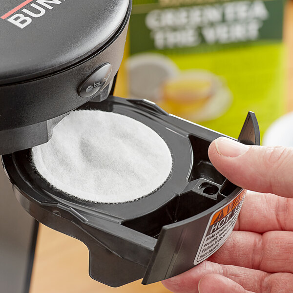 A hand using a black coffee maker to brew Bigelow Green Tea pods.