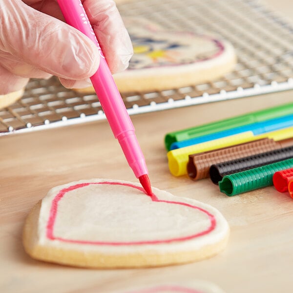 A hand with a glove using a pink Chefmaster food decorating pen to draw a heart on a cookie.