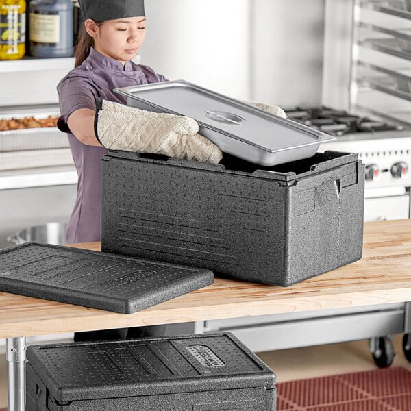 A woman in a chef's uniform putting a tray in a black CaterGator food pan carrier.
