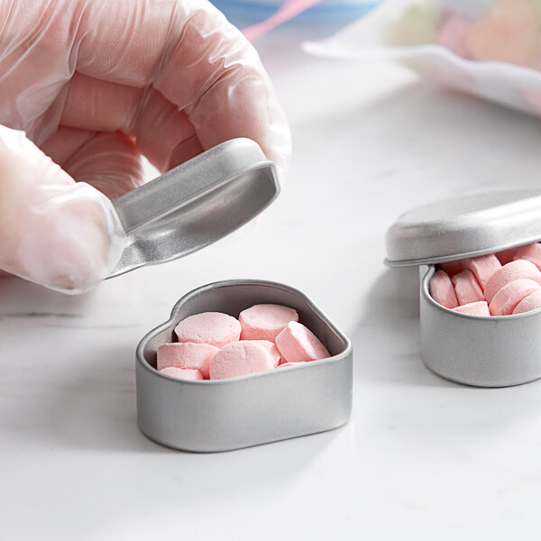 A hand holding a silver heart-shaped mini tin filled with pink candies.