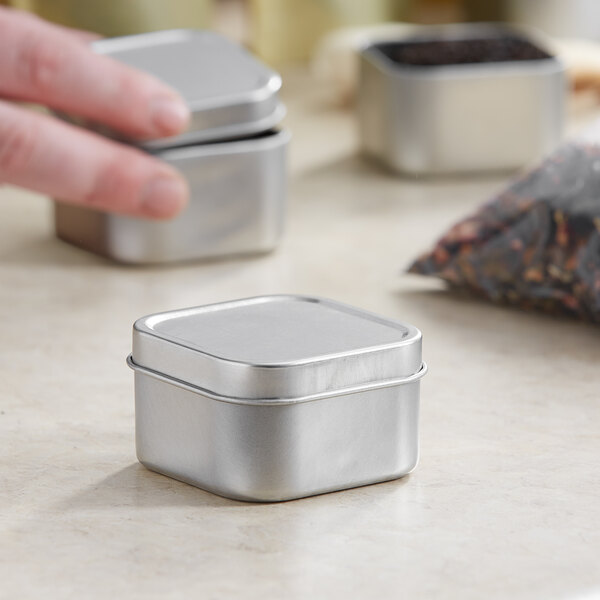 A hand holding a silver square metal tin with a slip cover on a counter.