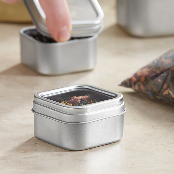 A person's hand pouring tea into a small open silver square tin with a clear window lid.