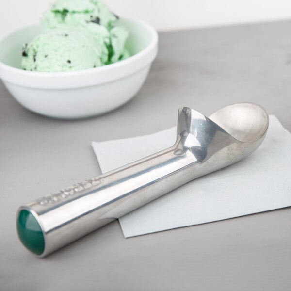 A Zeroll aluminum ice cream scoop with a green handle on a napkin next to a bowl of ice cream.