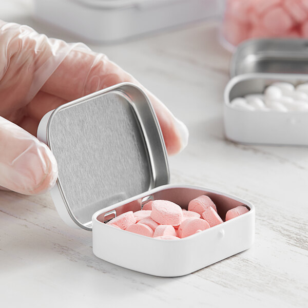 A hand holding a white rectangular metal tin filled with pink pills.
