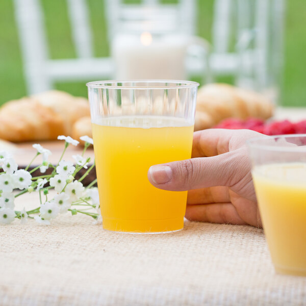 A person holding a WNA Comet Classicware clear plastic fluted tumbler filled with orange juice.