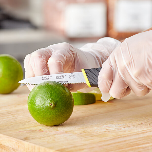 A hand in yellow gloves uses a Schraf serrated paring knife to cut a lime.
