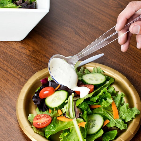 A hand holding a clear Fineline Platter Pleasers disposable ladle filled with white sauce over a bowl of salad.