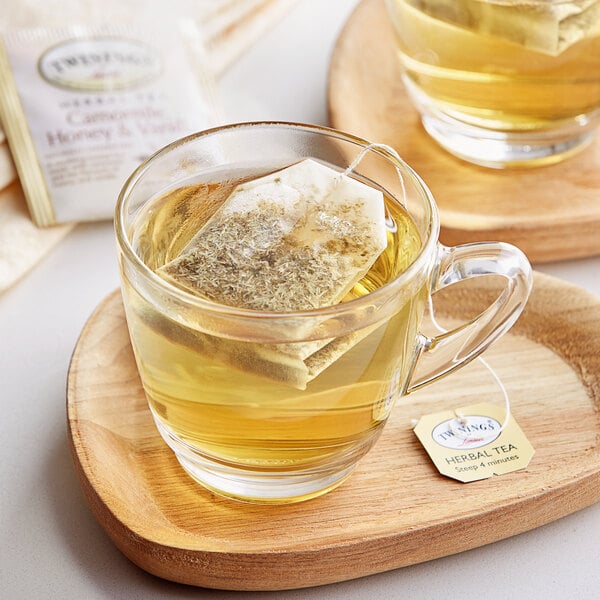 A glass mug of Twinings Chamomile, Honey & Vanilla tea with a tea bag in it on a wooden tray.