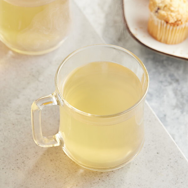 A clear glass mug of Twinings Lemon & Ginger tea next to a muffin.