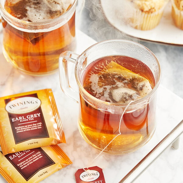 A glass mug of Twinings Earl Grey tea with a tea bag in it on a saucer with muffins.
