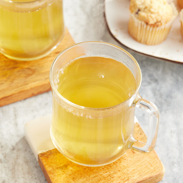 A wooden board with a plate of muffins and a glass mug of yellow Twinings Green Tea.