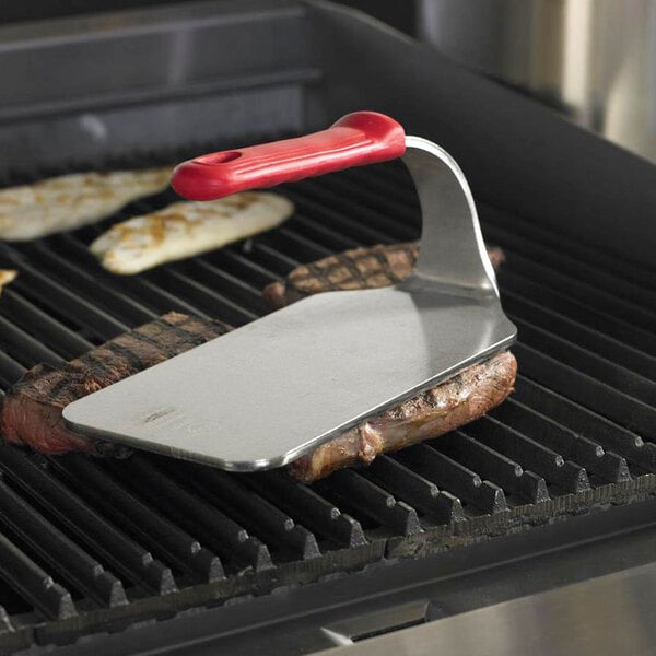 A Vollrath steak weight with a red silicone handle being used to cook a steak on a grill.