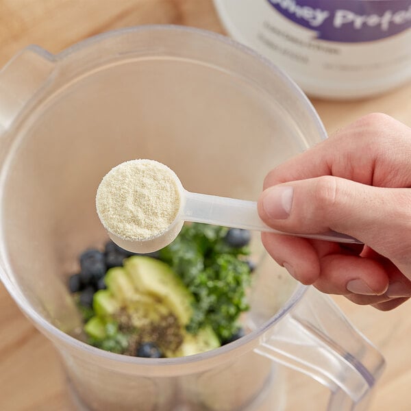 A person using a spoon to add Add A Scoop whey protein powder to a blender.