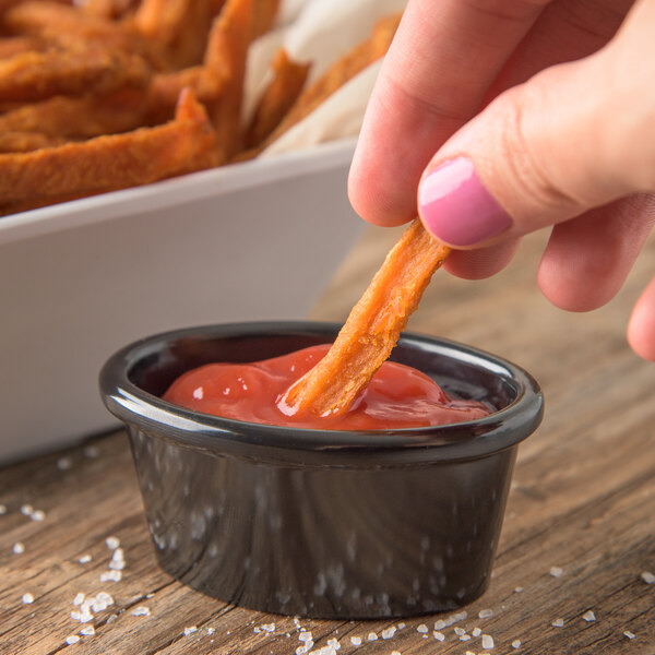 A person dipping a french fry into a Carlisle black melamine ramekin of ketchup.